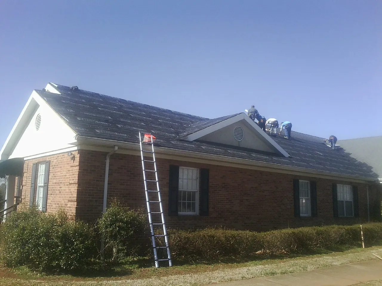 Crew tearing shingles off a brick ranch roof with a ladder set — Emergency Roof Repair work by our crew in Minong