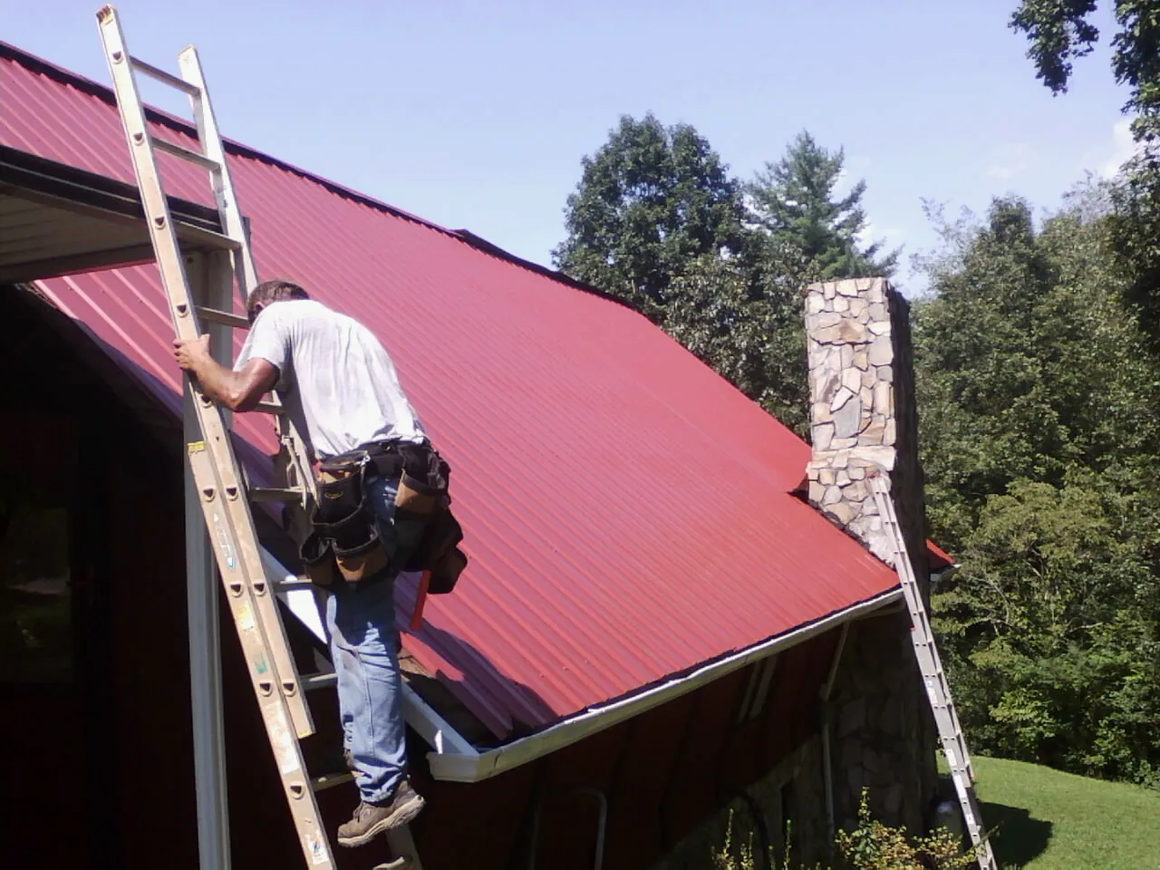 Roofer on a ladder beside a red metal roof and stone chimney — Soffit & Fascia Repair by Roofing Brothers in Minong